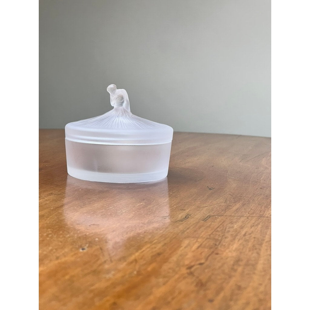 The Vintage Lalique France Ballerina Frosted Crystal Covered Powder Box by Lalique, featuring a sculpted ballerina lid, sits on a wooden surface against a plain, light background.