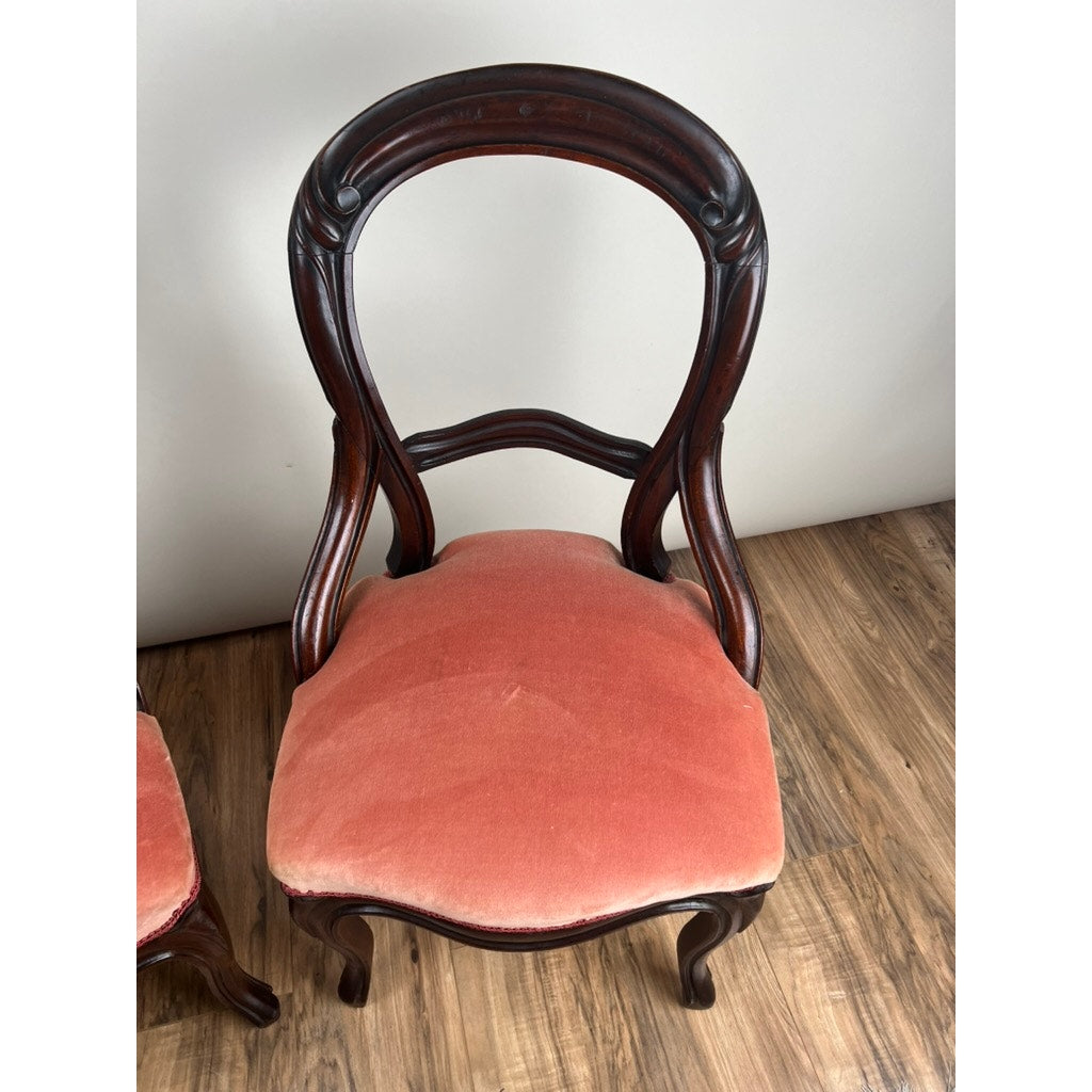 A pair of antique Victorian walnut balloon back side chairs with ornate curved backs and rose velvet upholstery sit on a wooden floor against a plain, light-colored wall. Brand: Unknown.
