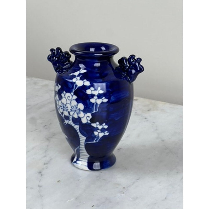 The Vintage Japanese Blue and White Floral Bud Vase features a deep blue hue, white floral designs, and two ornate handles. It sits on a white marble surface with a light gray background. Brand: None/Unknown.