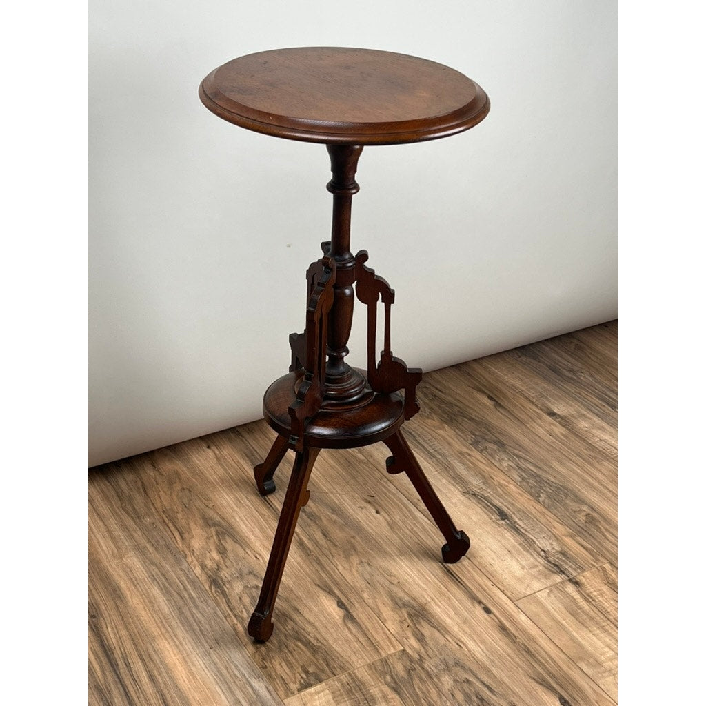 The Antique Victorian Walnut Candle Stand, Late 19th Century, features a small round top with a dark finish and stands on three intricately carved legs. It is showcased on a wood-patterned floor against a plain, light-colored wall.