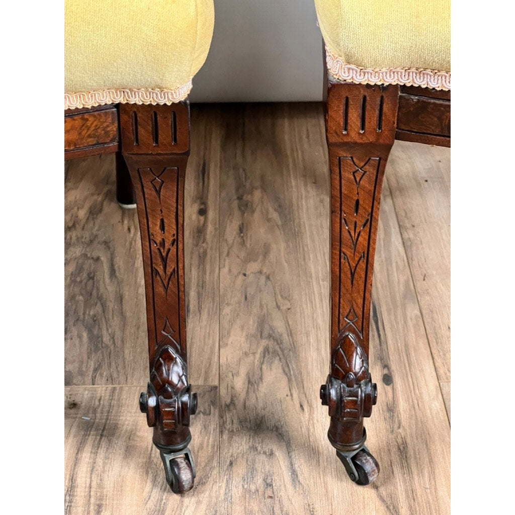 Close-up of two ornately carved walnut chair legs from a Fine Antique Pair Renaissance Revival Side Chairs, attributed to John Jelliff (Newark, c. 1880), showing intricate patterns, brass casters, and yellow upholstery on a wooden floor.