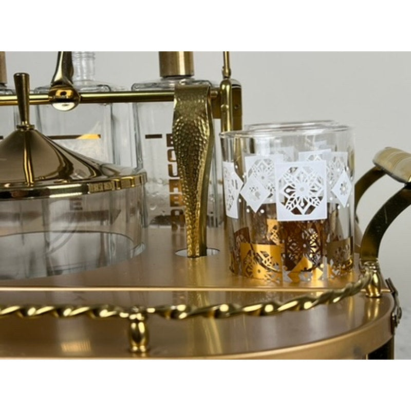 A close-up of a Vintage Mid Century Modern Bar Caddy c. 1960s displays a glass with white and gold geometric patterns, a gold cocktail shaker, and bottles in the background, creating an elegant retro atmosphere. Brand is unknown.