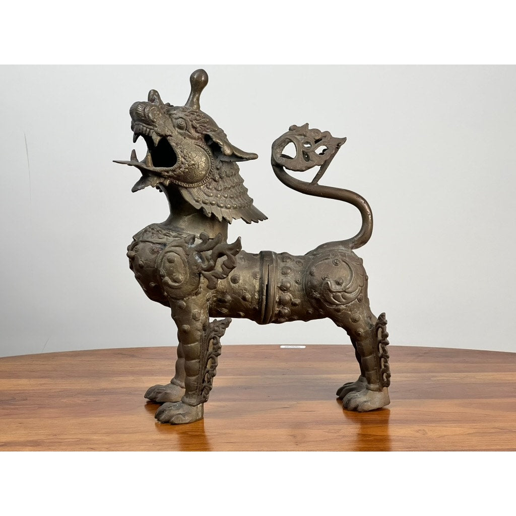 The Vintage 17” Cast Brass Pixiu Tibetan Lion c. 1960 is a detailed bronze statue of a mythical creature with a curled tail, shown on wood against a plain background—an elegant piece reflecting Asian decorative arts. Brand: None/Unknown.