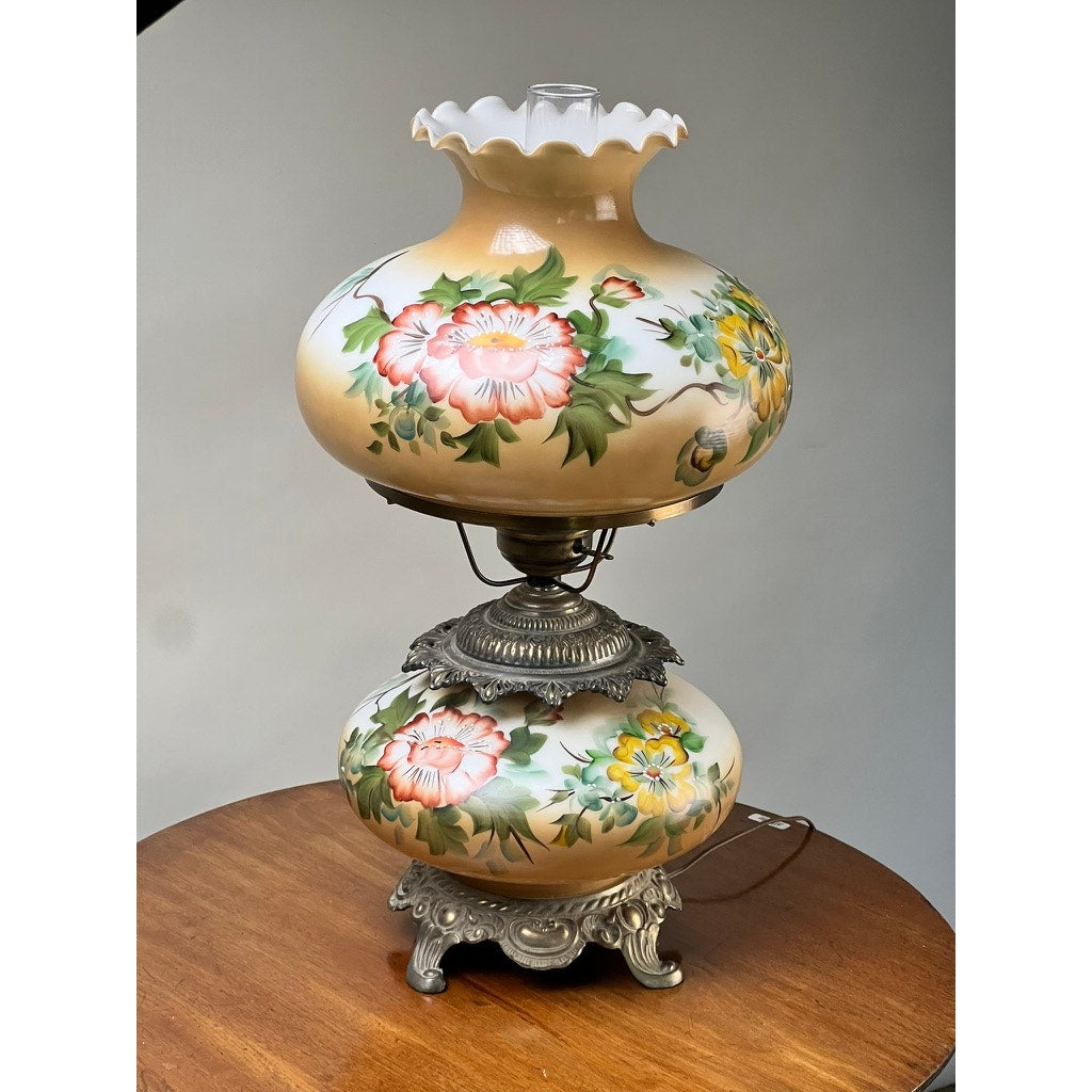 A Vintage Hand-Painted Floral Milk Glass Hurricane Parlor Lamp with ornate metal base and pink, yellow, and green floral designs on the glass shade and base is displayed on a wooden surface against a plain background. Brand: Unknown.