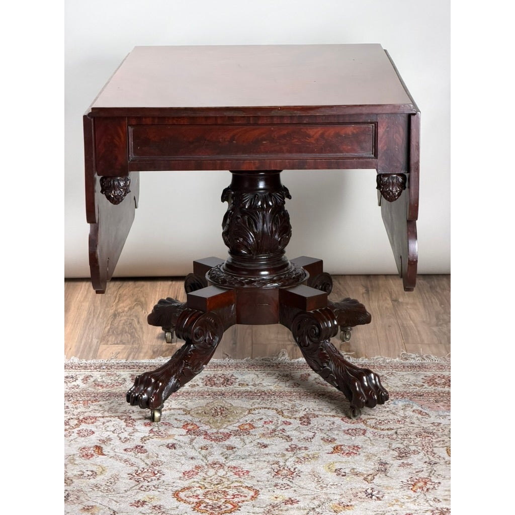 American Classical Carved Mahogany Drop-Leaf Breakfast Table, c. 1825–1840, featuring carved acanthus leaf details, four carved legs, and claw-and-ball feet on casters.