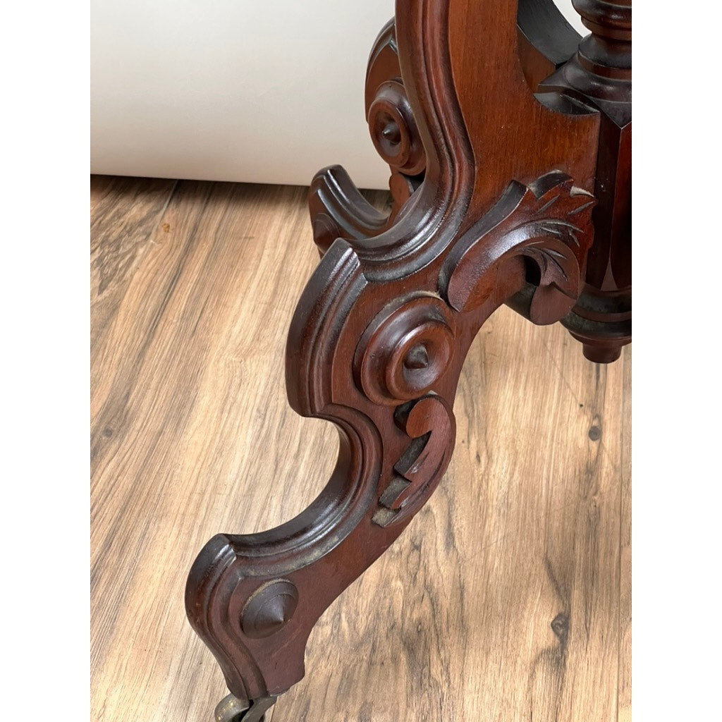 An ornate, dark wood leg from an Antique Victorian Walnut Lamp Stand (late 19th century, maker unknown) shows intricate carvings and small wheels, resting on a light wood floor with a blurred white surface in the background.