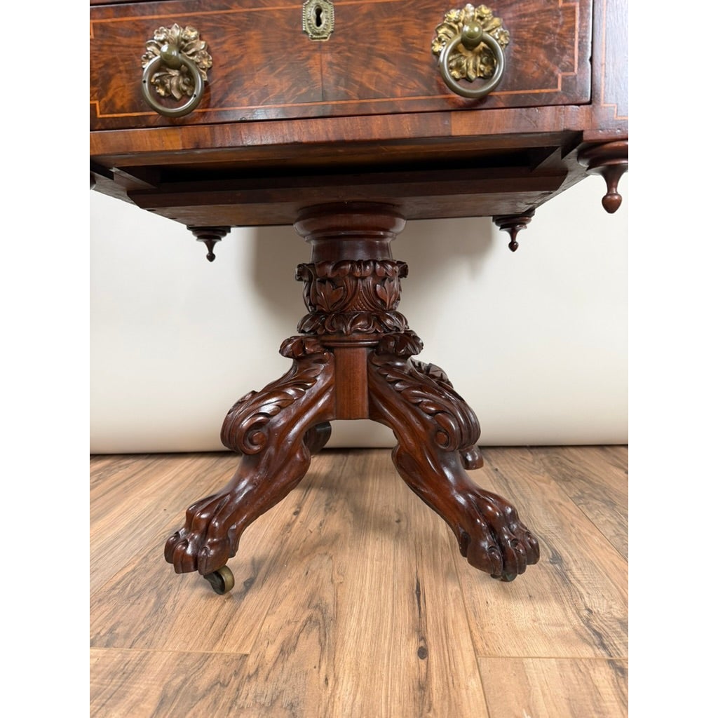 An American Classical Figured Mahogany Work Table, circa 1830–1845, featuring a single drawer with brass ring pulls, a central pedestal column with carved foliage, and tripod claw-and-ball feet with casters.