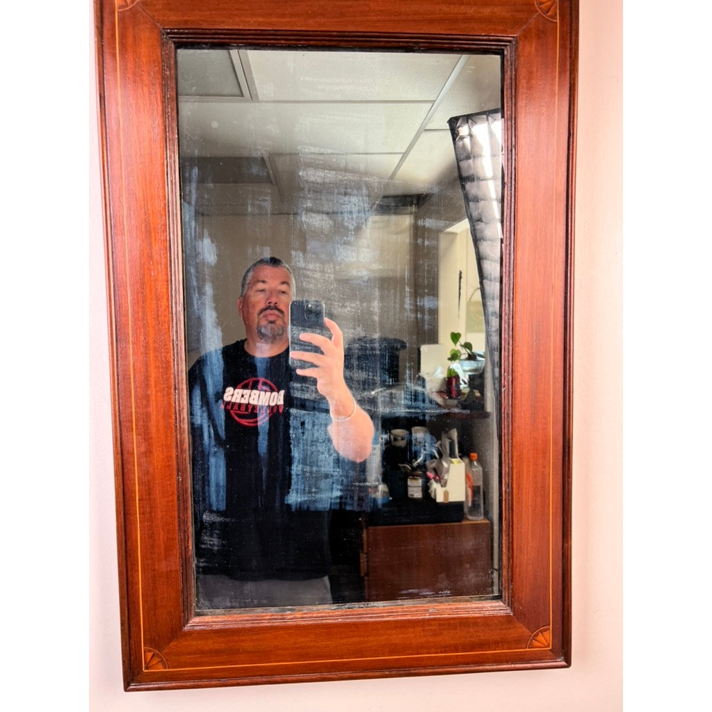 A man takes a mirror selfie in an American Federal Mahogany Wall Mirror with Fan Inlay, featuring its original hand silvered glass from circa 1810–1825. The glass is streaked and smudged, and the room behind him contains cabinets, shelves, a plant, and various items.