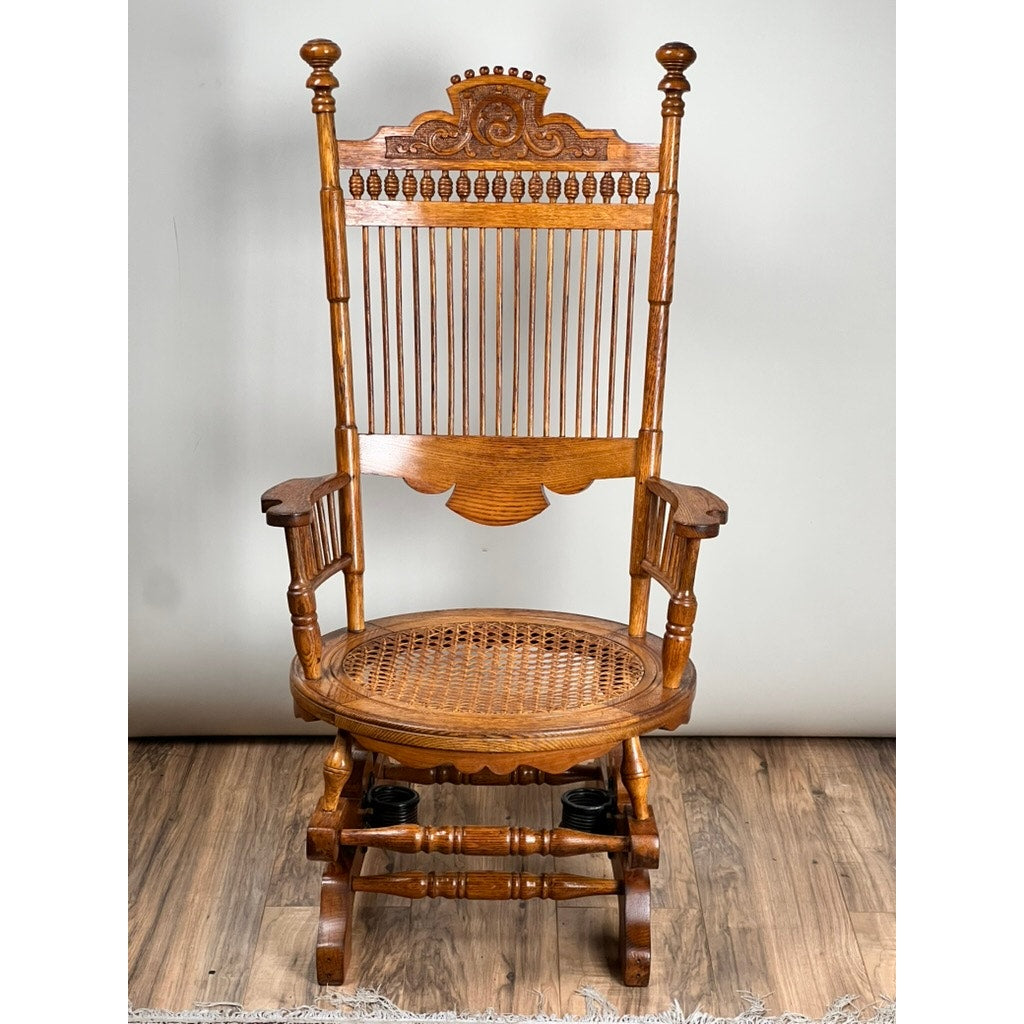 The Antique Victorian Oak Platform Rocker c. Late 19th Century features a high, ornate backrest, spindle detailing, and a woven cane seat—an eye-catching vintage piece set against a light-colored wall on hardwood flooring.