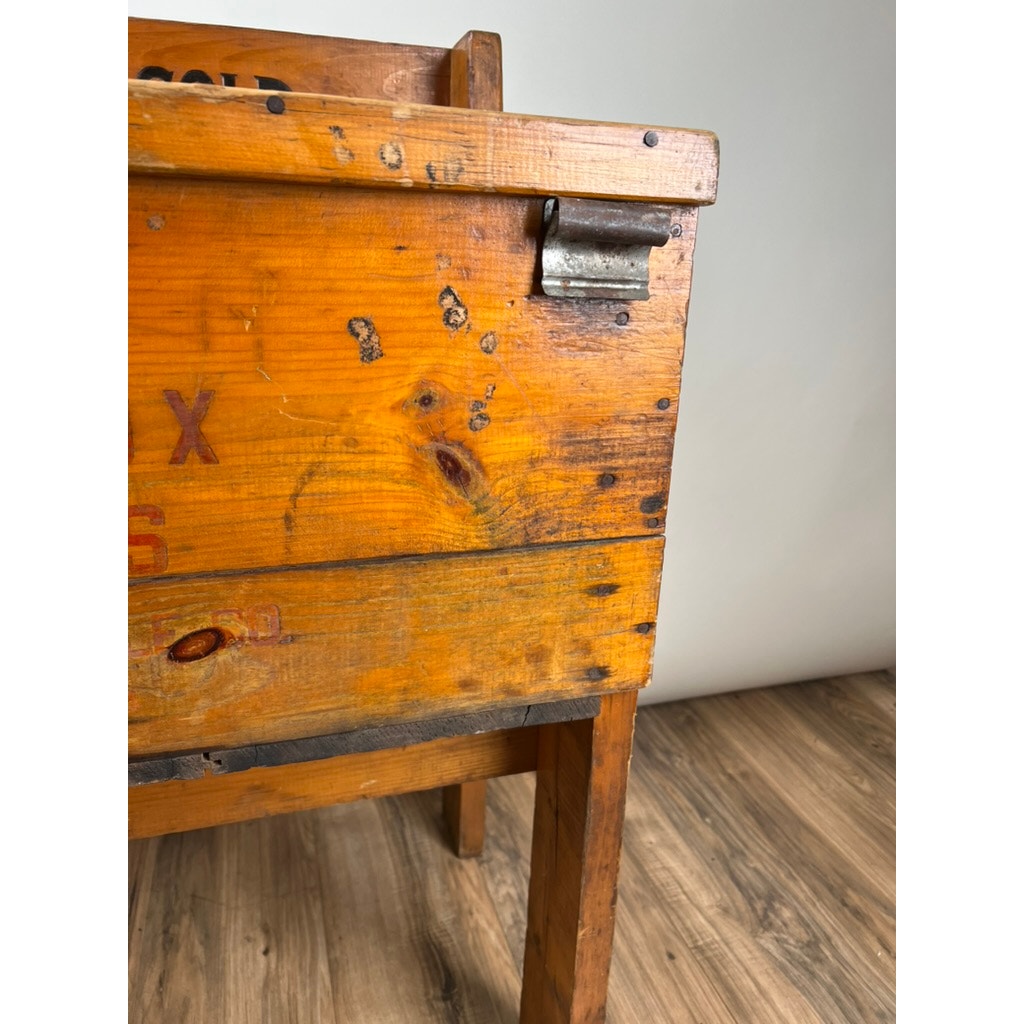 Close-up of a worn Antique Tin Lined Pine General Store Red Fox Ginger Ale of Rhode Island Cooler c. 1930, featuring orange-stained wood, metal hardware, faded writing, and scratches, sitting on a wood floor against a plain background.