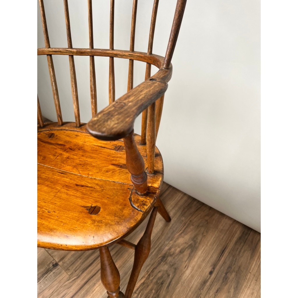 A close-up of an Antique Near Pair 18th Century Windsor Chair, featuring a spindle back and armrest, set on a wooden floor against a light wall—showcasing the craftsmanship of early American furniture. Brand: Unknown.