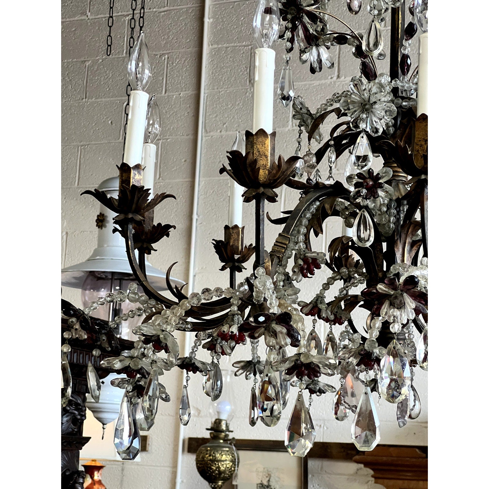 A close-up of an Antique Late 19th Century French Rococo Tole Amethyst and Crystal Ten Light Chandelier displays candle-style lights, crystal prisms, and ornate metalwork, with antique lamps and framed art against a light brick wall in the background.