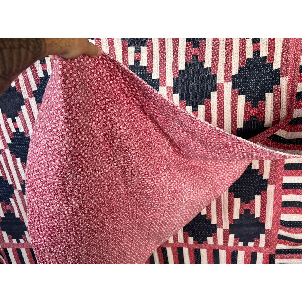 A hand lifts the corner of a pink and white textile, revealing its underside. Behind it lies a Matching Pair of Antique American Patriotic Hand Quilted Log Cabin Quilts c. Early 20th Century, with red, white, and black geometric patterns.