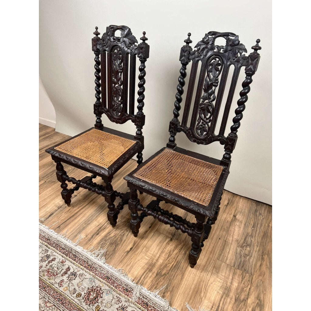 A Pair of Antique French Walnut Hunting Chairs c. Late 19th Century, featuring tall carved backs, twisted legs, and caned seats, are side by side on a wooden floor near a patterned rug and plain light wall.