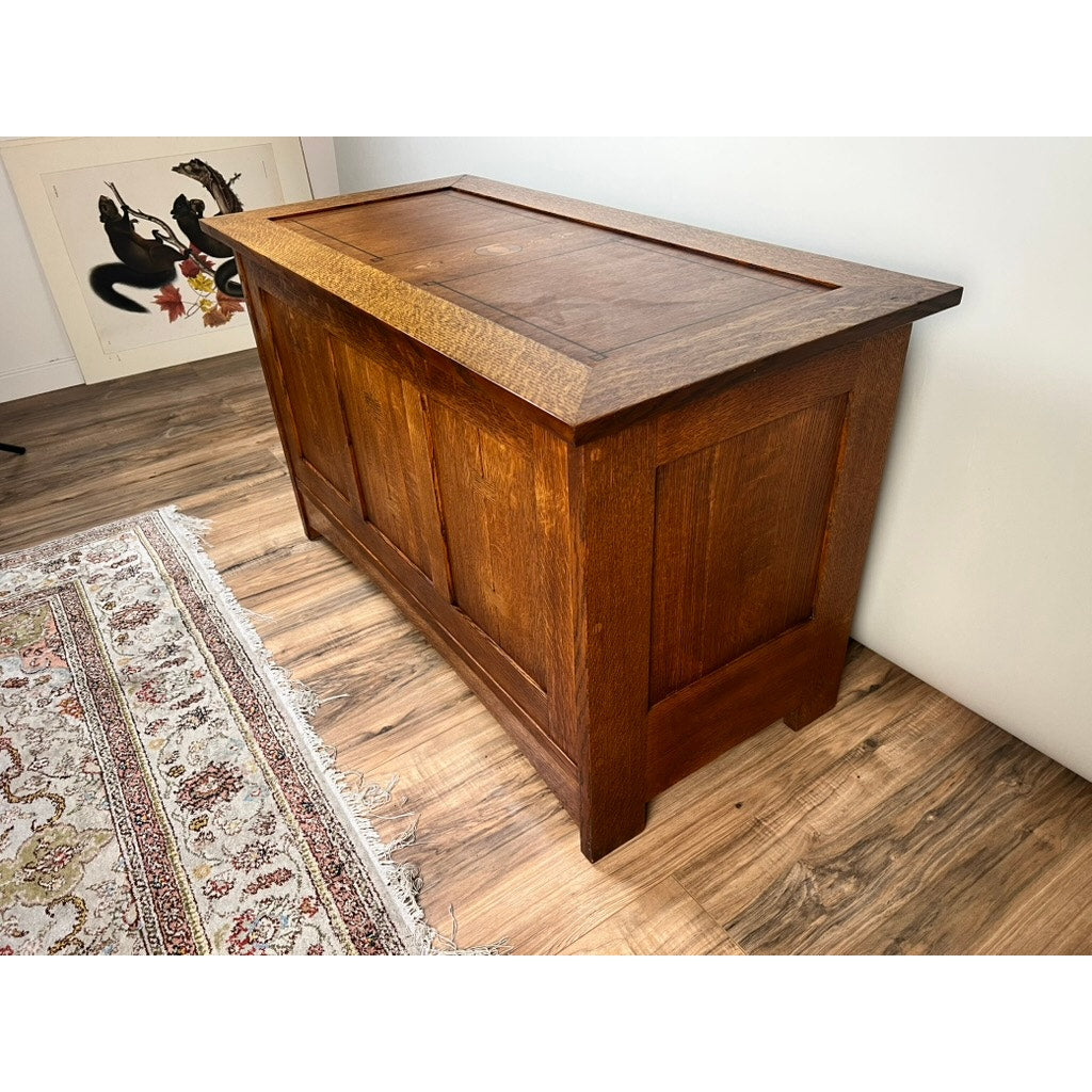 A Stickley Vintage Harvey Ellis Millennium Edition Oak Blanket Chest with paneled sides and a rectangular lid sits on a wood floor by a patterned rug. A framed silhouette artwork leans nearby, evoking classic Arts & Crafts style.