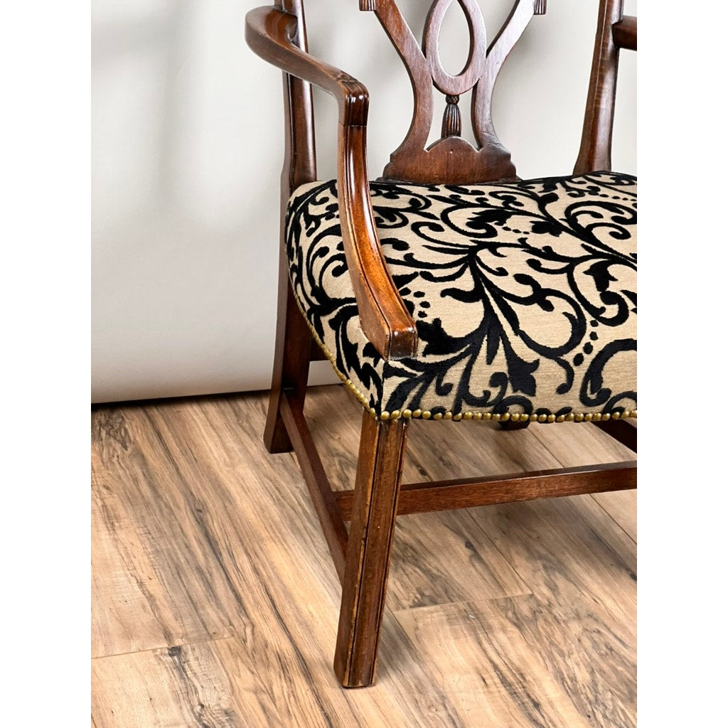 A close-up of an Antique George III Chippendale Open Arm Chair (early 19th century) with curved arms, a decorative backrest, and a black ornate floral patterned upholstered seat on beige, set on a light wood floor—an elegant English antique.