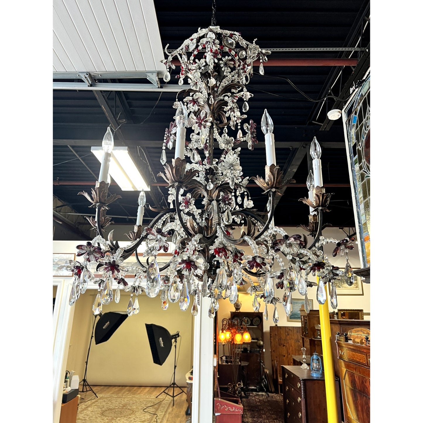 An Antique Late 19th Century French Rococo Tole Amethyst and Crystal Ten Light Chandelier hangs from the ceiling in a room featuring studio lights, vintage furniture, and a stained glass window in the background.