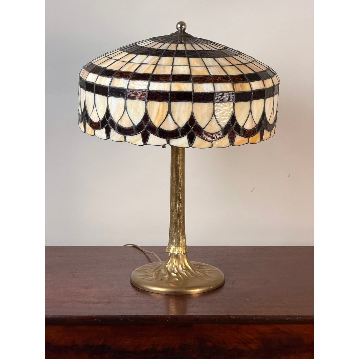 A Pearl City Antiques Vintage Tiffany Revival Leaded Stained Glass Table Lamp with a cast brass tree-form base and a cream, brown, and black patterned shade rests gracefully on a wooden surface.