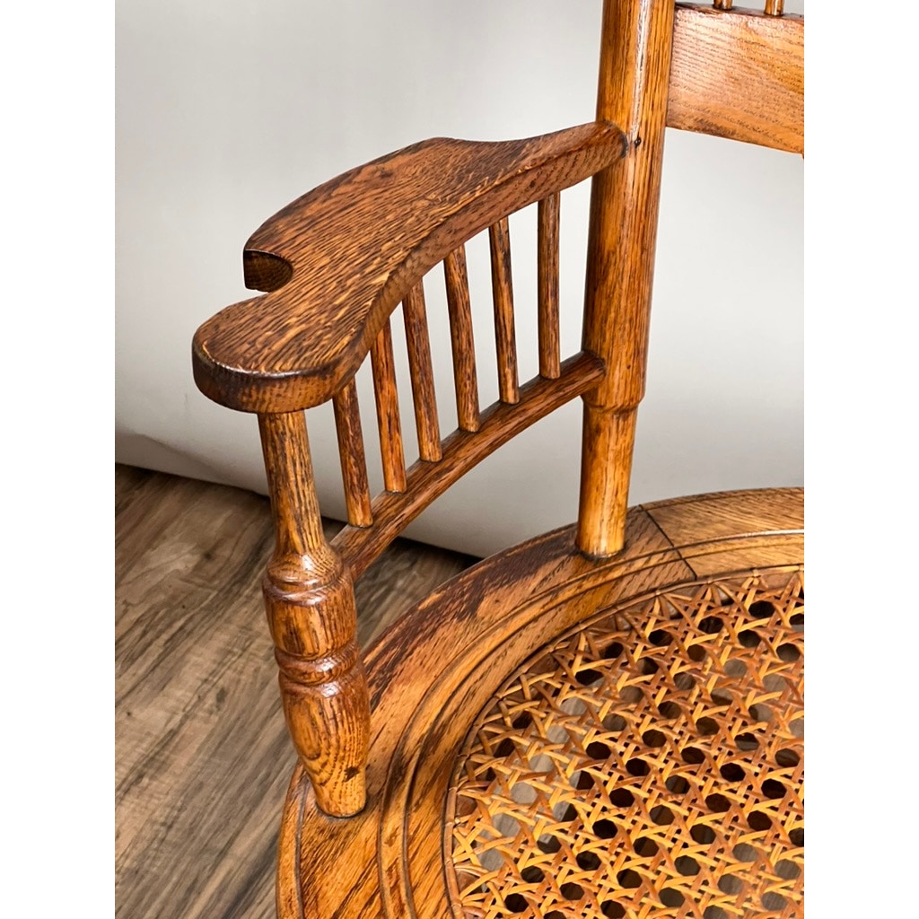 Close-up of an Antique Victorian Oak Platform Rocker c. Late 19th Century featuring vertical spindle armrests, a round woven cane seat, and a polished finish. The piece is placed on a wooden floor with a neutral backdrop. Brand: Unknown.