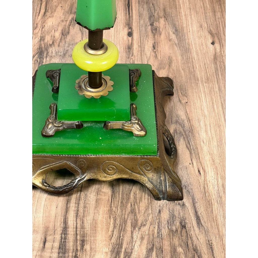A close-up of the ornate base of an Antique Art Deco Uranium and Slag Glass Lamp c. 1920, featuring green and brass detailing, resting on a wood-grain surface. Only the bottom of the lamp is visible; brand unknown.