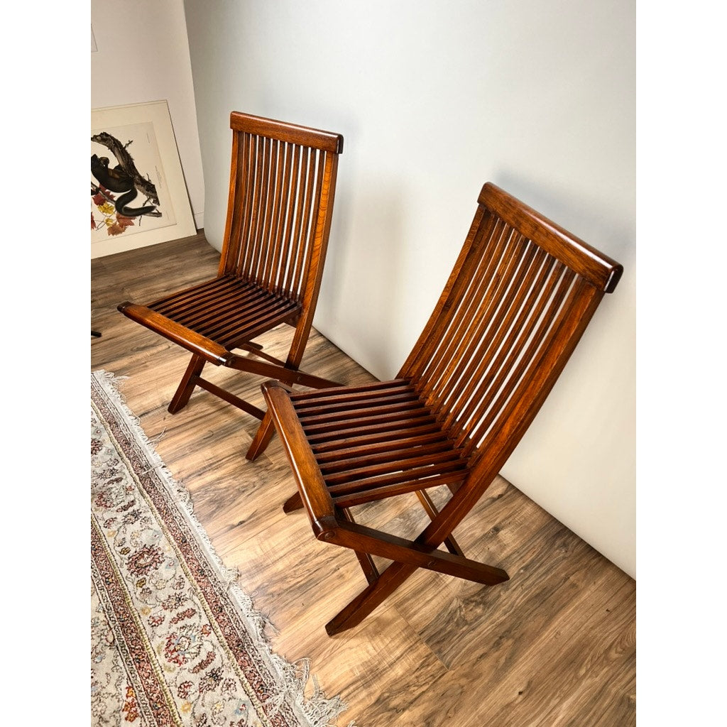 A Pair of Vintage Mid-Century Modern Style Teak Folding Chairs sit side by side on a wood floor near a patterned rug, with framed artwork leaning against the wall in the background. Brand: None/Unknown.
