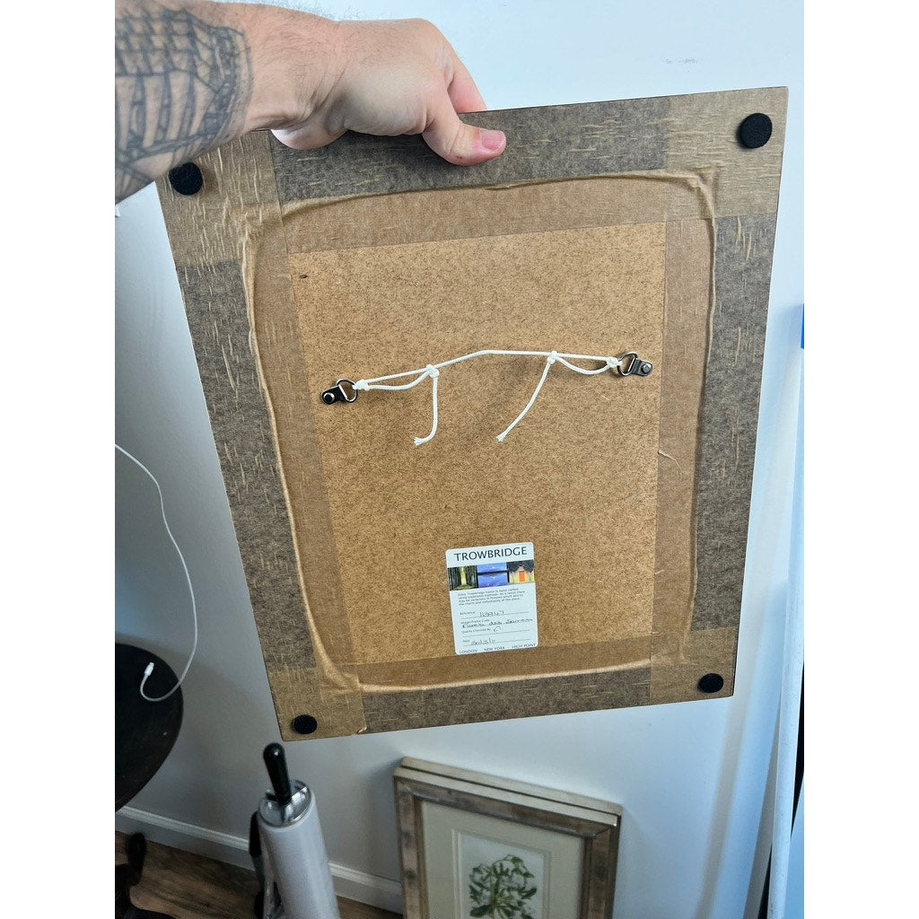 A tattooed hand holds the back of a Vintage Trowbridge Custom Framed Botanical Print “Gladiolus × willmoreanus” after Louis van Houtte, showing its hanging wire and label, while another vintage botanical print rests on the floor below.