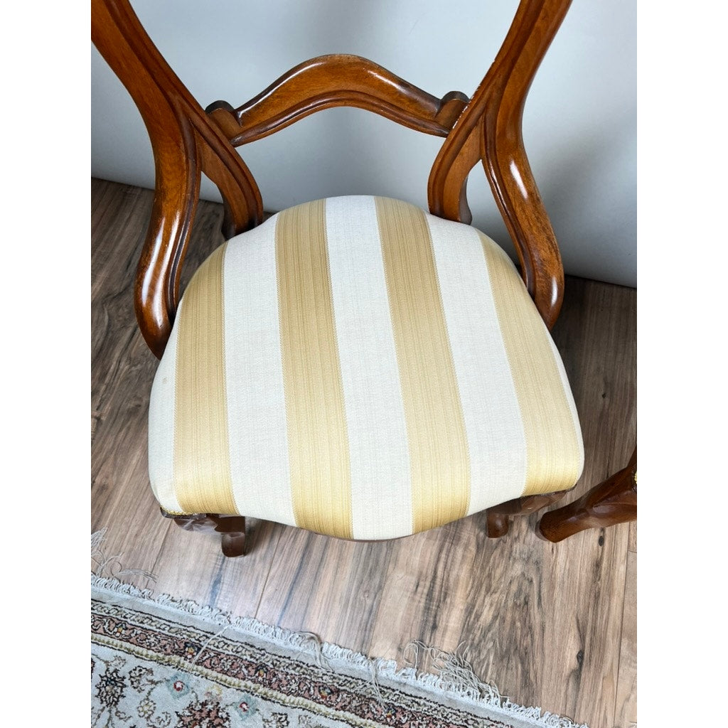 A pair of antique Victorian balloon back chairs from the late 19th century, featuring curved backrests and cushioned seats with beige and yellow striped upholstery, sit on a wood floor beside a patterned rug.