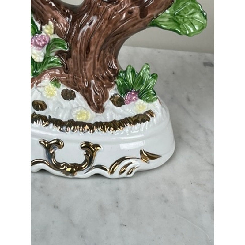 A close-up of a Vintage Lena Liu Porcelain Hummingbird Accent Lamp, featuring a brown tree trunk, green leaves, and pastel flowers on a white base with gold accents, displayed on a marble surface.
