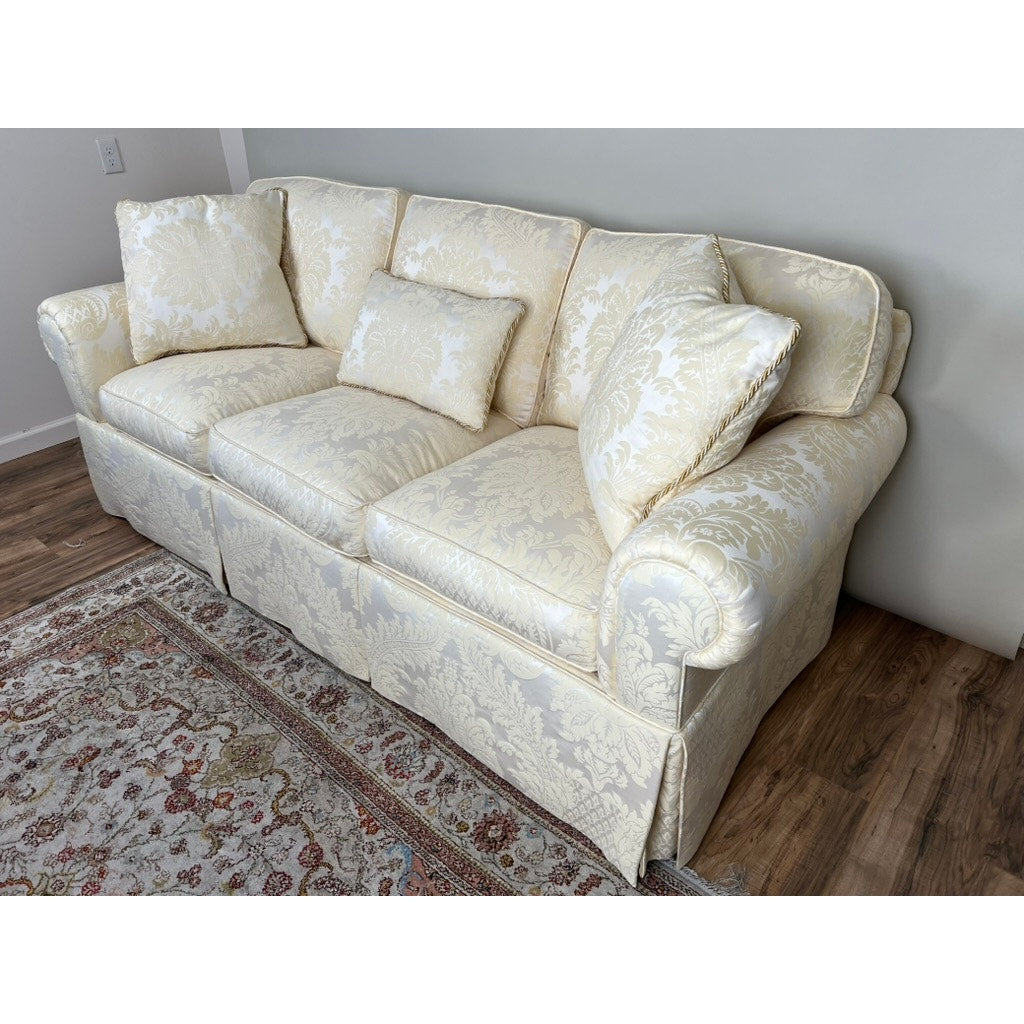 A Vintage Custom Ivory Upholstered Sofa by Stickley with three seat cushions and four matching throw pillows sits on a wooden floor beside a patterned area rug and a light-colored wall.