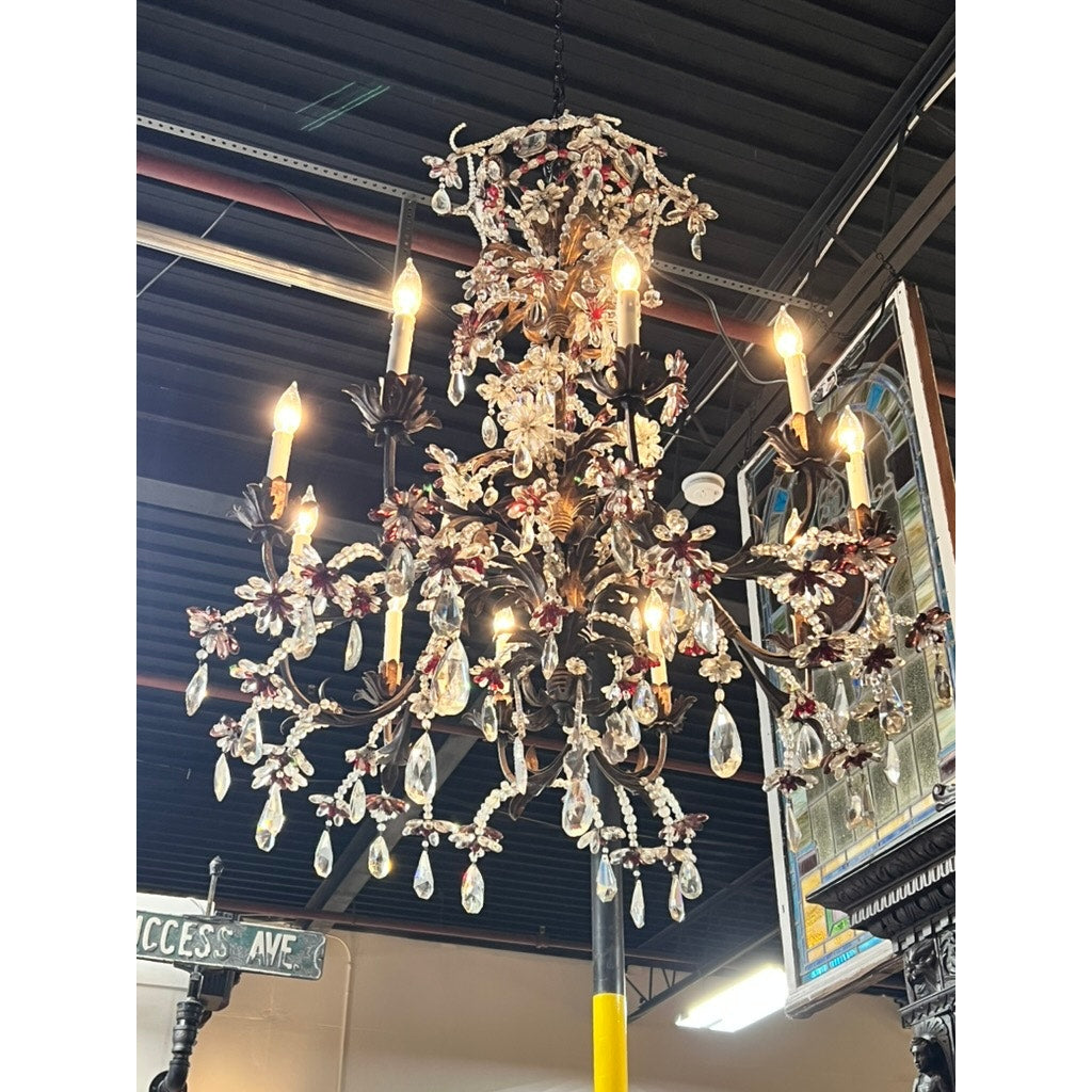 An Antique Late 19th Century French Rococo Tole Amethyst and Crystal Ten Light Chandelier with lit bulbs, crystal prisms, and red glass accents hangs from a dark ceiling near a street sign and stained glass window. Brand: Unknown.