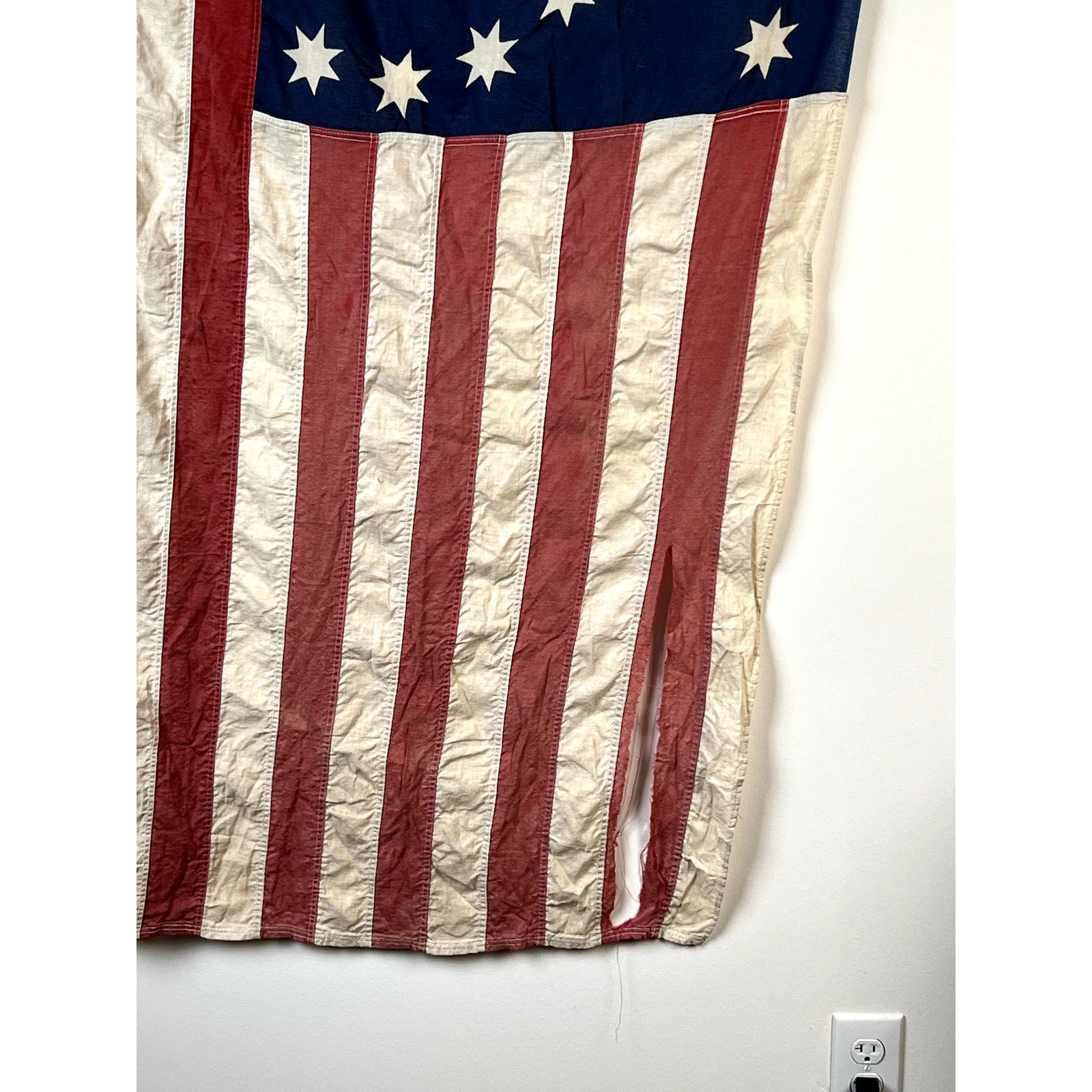 A Vintage Valley Forge “76” Bennington American Flag from the Bicentennial Era (c. 1976), featuring a large vertical tear in the lower right corner, hangs on a white wall above an electrical outlet.