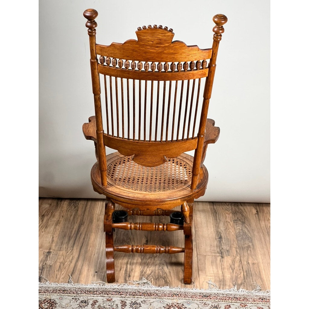 The Antique Victorian Oak Platform Rocker (Late 19th Century) features a high spindle back, decorative carvings, curved arms, and a round cane seat, evoking classic charm on a wooden floor with part of a patterned rug visible.