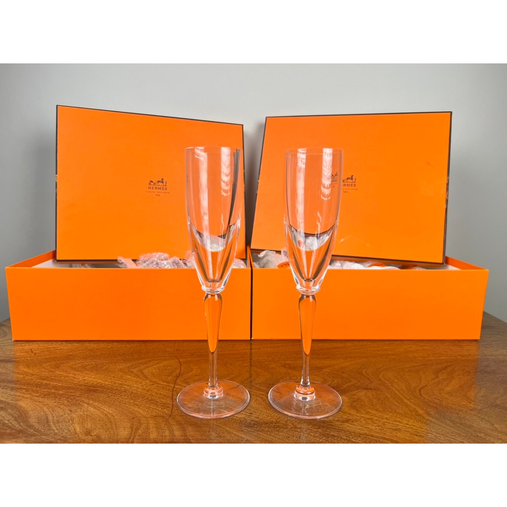 A set of eight Hermès Crystal Champagne Flutes by Saint Louis France (10.5") is displayed on a wooden surface with two open orange Hermès boxes and white tissue paper, set against a plain, light background to highlight this luxury glassware.