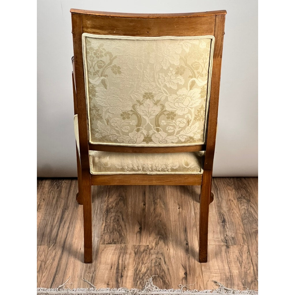 An Antique French Directoire Style Fauteuil Open Arm Chair in Sapelle Mahogany (late 19th c.) features a light beige floral upholstered backrest and seat. Seen from the back on a wood floor, it displays timeless elegance against a plain wall.
