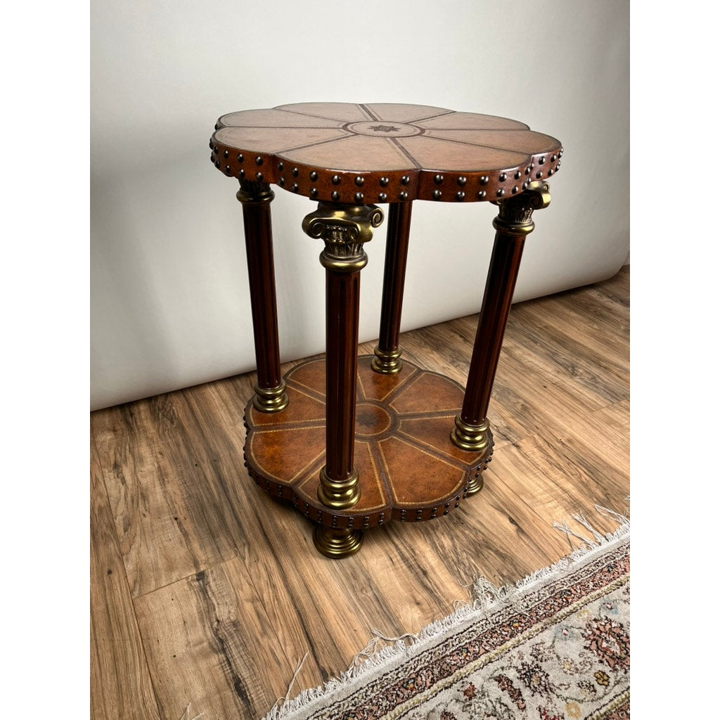 The Vintage Maitland-Smith Leather-Clad Occasional Table by Maitland Smith features a flower-shaped top, brass nailhead trim, ornate brass accents on six columns, and a matching lower shelf. It is shown on a wooden floor beside a patterned rug.