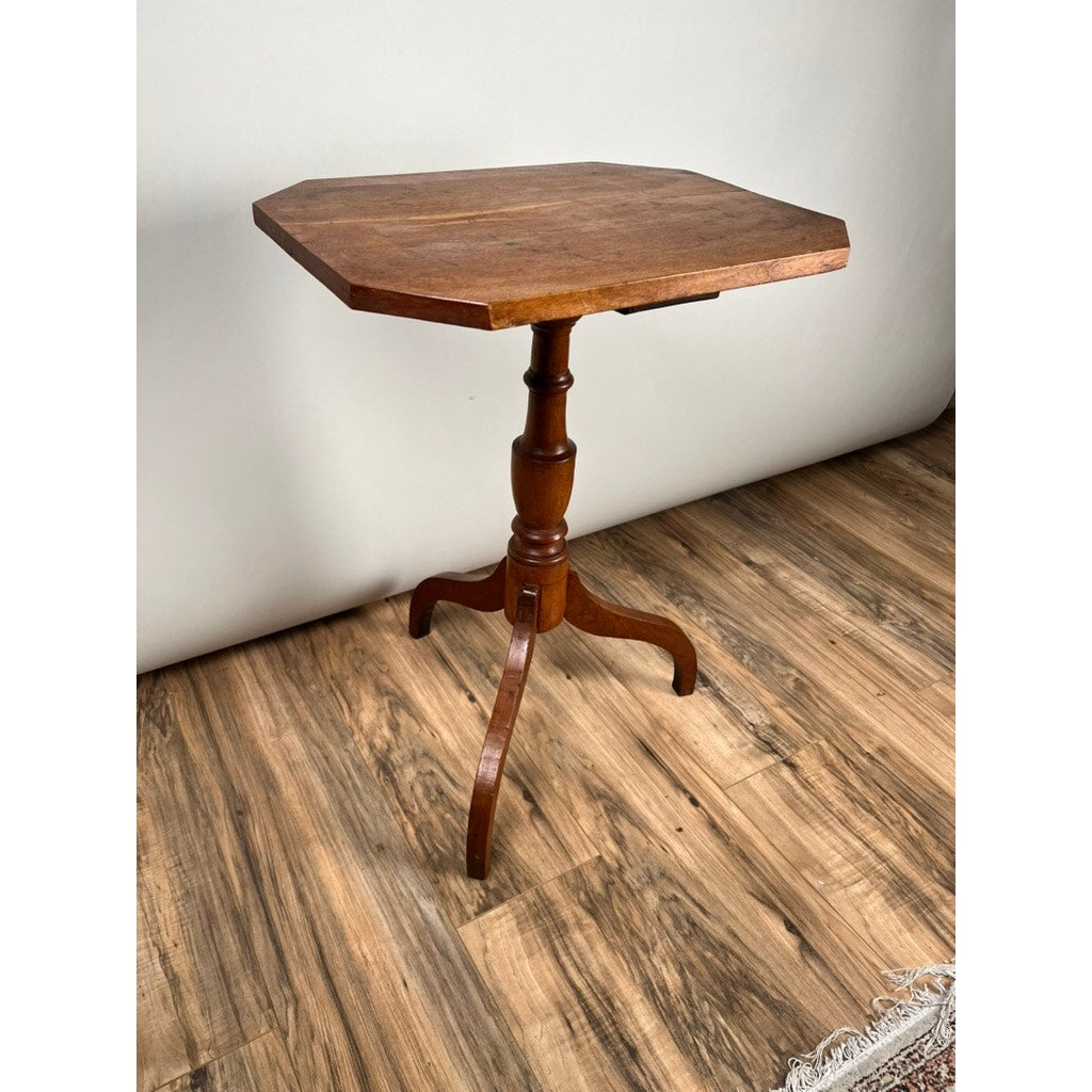 An Antique 19th Century Cut Corner Hepplewhite Candle Stand with an octagonal top, central turned column, and three curved legs stands on a wooden floor near a white wall and the edge of a patterned rug.