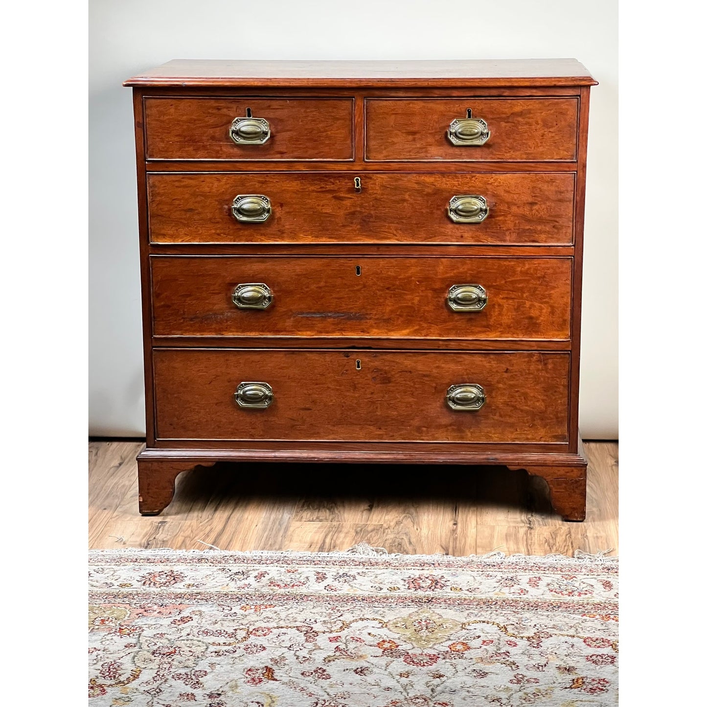 A Late Georgian English Mahogany Chest of Drawers, circa 1780–1810, with two smaller top drawers and three larger lower drawers featuring brass handles, stands on a patterned rug against a white wall.