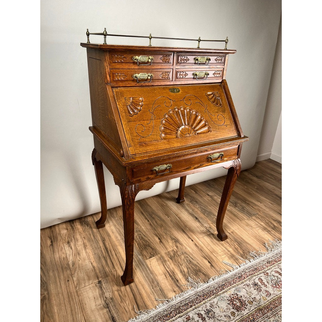 The Antique Victorian Oak Ladies Writing Desk c. 1900 features ornate carvings, brass handles, four slender legs, three small drawers, and a slanted floral front panel. Brand unknown. It stands on a wood floor next to a patterned rug.