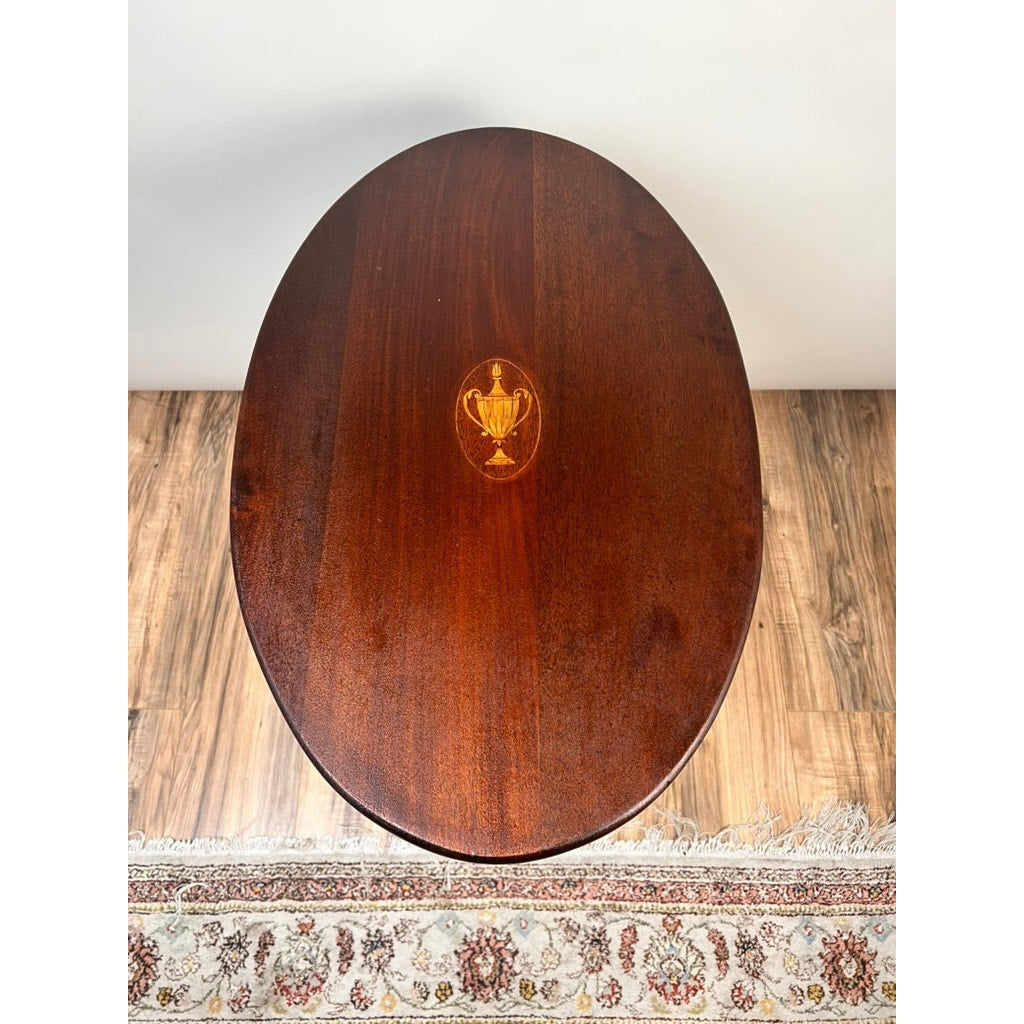 Antique George III Tilt-top Tea Table c. Early 19th Century by Pearl City Antiques—an elegant oval wooden table with a polished finish and a distinctive small golden urn inlay at its center. Reminiscent of classic tea tables, this piece sits gracefully on a wood floor near the edge of a decorative, fringed rug.
