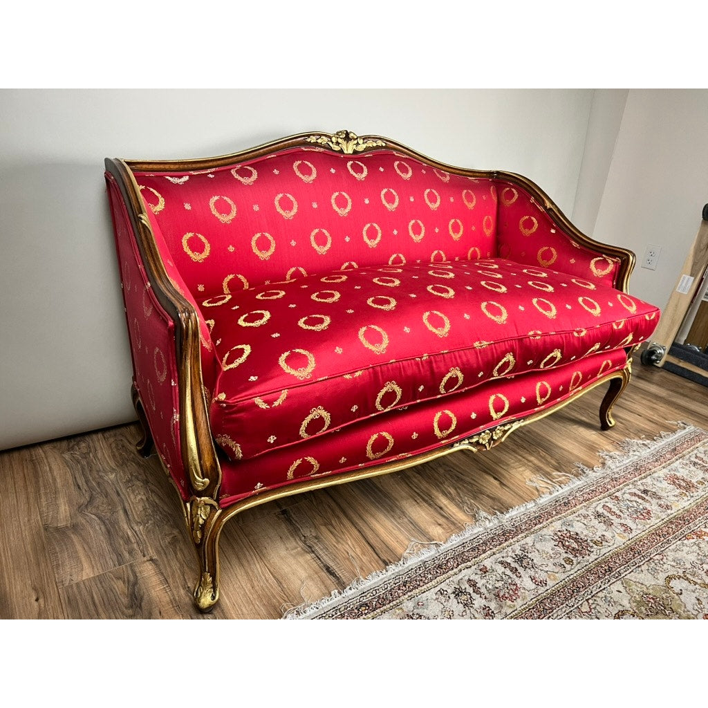 A Vintage French Regency Style Custom Upholstered Settee with a giltwood frame and red and gold upholstery sits on a hardwood floor beside a patterned rug, placed against a plain light-colored wall.