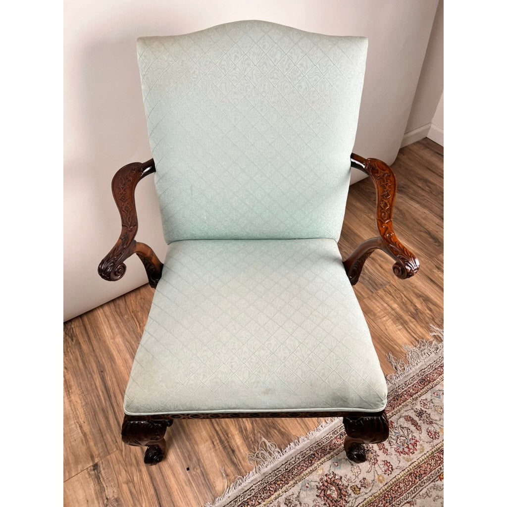 An Antique George II Mahogany Gainsborough Open Arm Chair (c. Mid 19th Century) with curved armrests, cabriole legs, and intricate carvings features light blue patterned upholstery and rests on a wooden floor by a decorative rug.