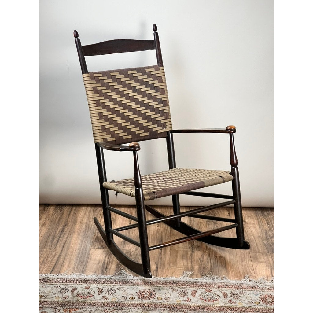The Antique Shaker #6 Tape Seat Rocker by Mt. Lebanon Shakers (late 19th c.) with a brown and beige geometric woven seat and backrest rests on a wood floor, partially bordered by a patterned rug in front.