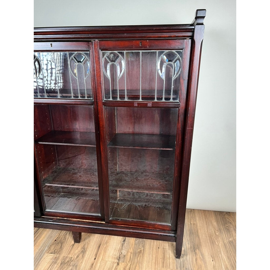 An Antique Arts & Crafts Leaded Glass Mahogany Bookcase c. 1920 stands on wooden flooring against a plain light wall, featuring glass doors, decorative leaded glass panels at the top, and empty shelves. Brand unknown.