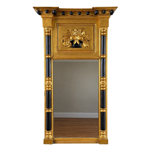 The Massachusetts Federal Giltwood & Ebonized Pier Mirror with Fruit Basket, c.1820 from Pearl City Antiques features a tall giltwood frame with ebonized (black) accents, decorative knobs, and a floral relief design at the top. It stands elegantly against a plain wall and wooden floor.