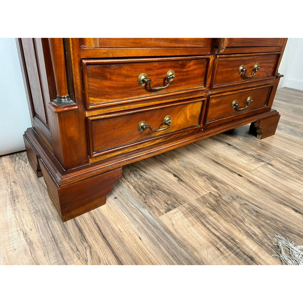 A wooden dresser with brass handles and four drawers stands on a wood-patterned floor beside a Vintage Victorian Style Glass Door Mahogany Bookcase. The dresser features sturdy legs, a polished finish, and a warm brown tone.