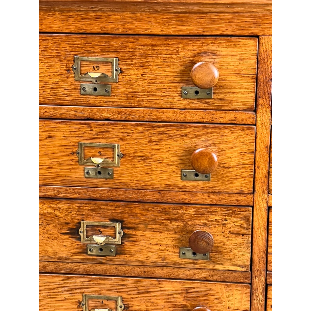 Close-up of an Antique Early 20th Century Oak Multidrawer File with Roll Top Pigeon Hole Top, featuring multiple drawers with metal label holders and round knobs. The oak wood displays a warm, polished finish and visible grain patterns. Brand: Unknown.