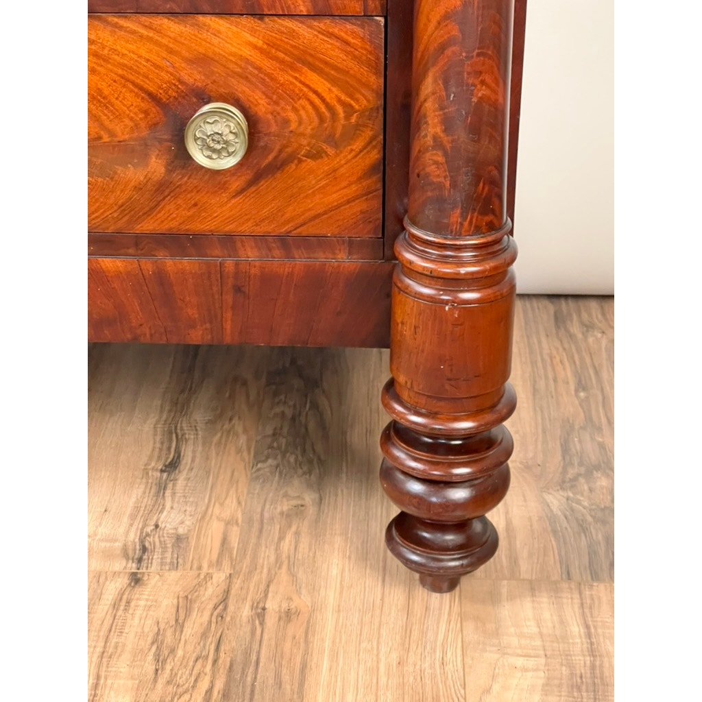 A close-up of an ornate, rounded leg on the Antique American Empire Albany Four Drawer Chest c. 1840, featuring a reddish-brown flame mahogany finish and brass drawer pull, standing on a wood-patterned floor. Brand: unknown.