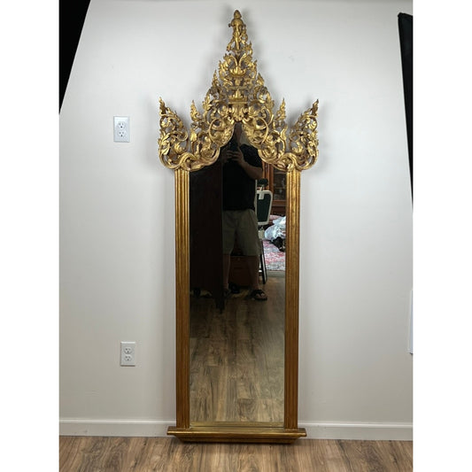 An Antique Giltwood Burmese Shrine Mirror c. 19th Century from Pearl City Antiques, featuring a tall rectangular shape with an ornate gilt frame and intricate pointed top, stands against the wall. Its reflection captures part of the room and a person in dark clothing holding a phone.