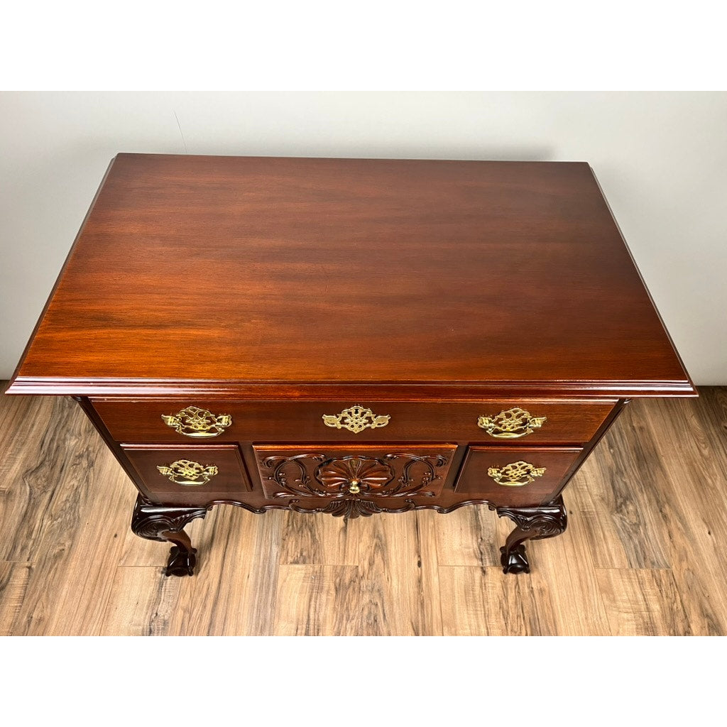 A Vintage Solid Mahogany Chippendale Style Lowboy by Councill Craftsmen, featuring ornate brass handles, decorative carvings, three drawers, and cabriole legs, is placed on a wood floor against a plain wall.