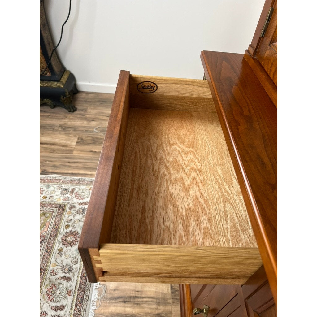 A partially open empty drawer is seen in a Vintage William IV Style Cherry Glass Door Bookcase by Stickley. A patterned rug covers the wood floor, with other furniture and a wall visible in the background. The drawer's interior is light wood.